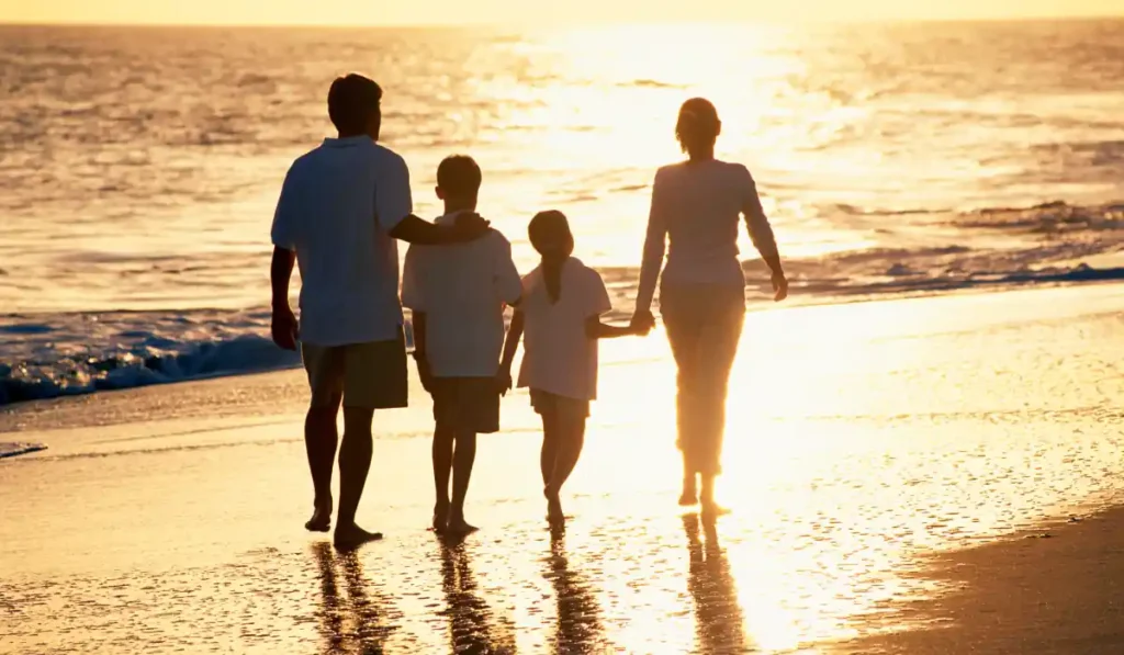 A family of four is walking along the beach at sunset. The sun is setting over the ocean, casting a warm golden glow on the sand. The family members are holding hands and enjoying a peaceful stroll, silhouetted against the bright sky.
