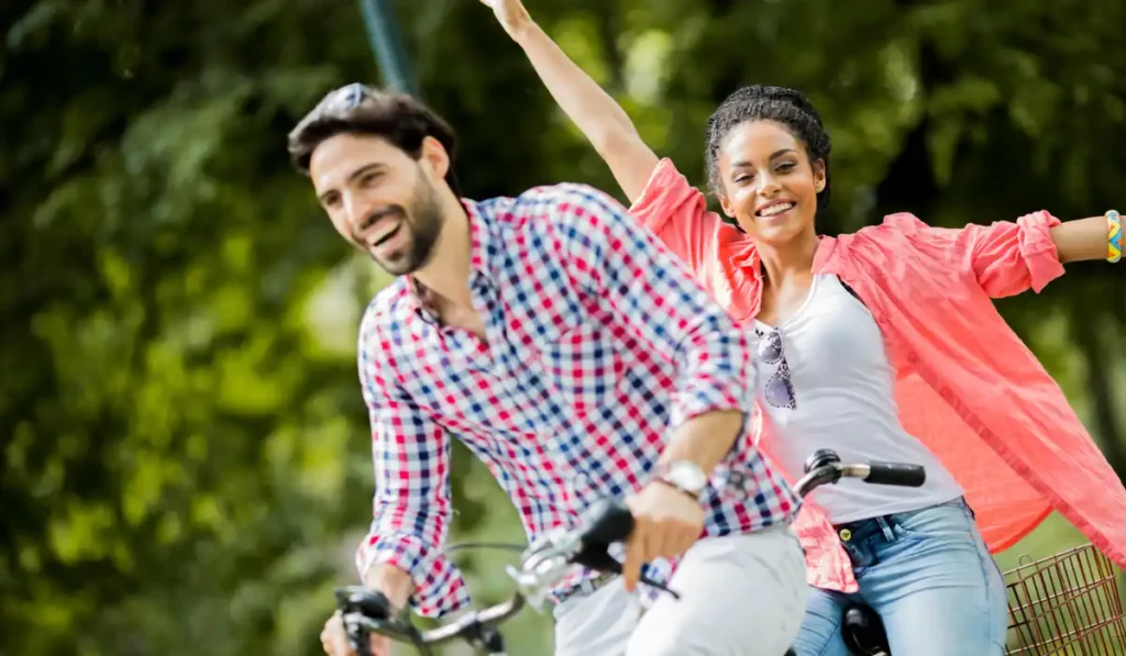 A man and a woman are joyfully riding bicycles outdoors. The man, wearing a checkered shirt, is in the foreground, while the woman in a pink shirt raises her hands in excitement. The background is lush and green, suggesting a park setting.
