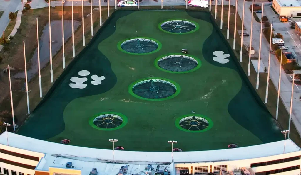 Aerial view of a golf driving range with large target circles on a turf field. Six white sand traps are visible on both sides. Surrounding the range are buildings and parking areas.
