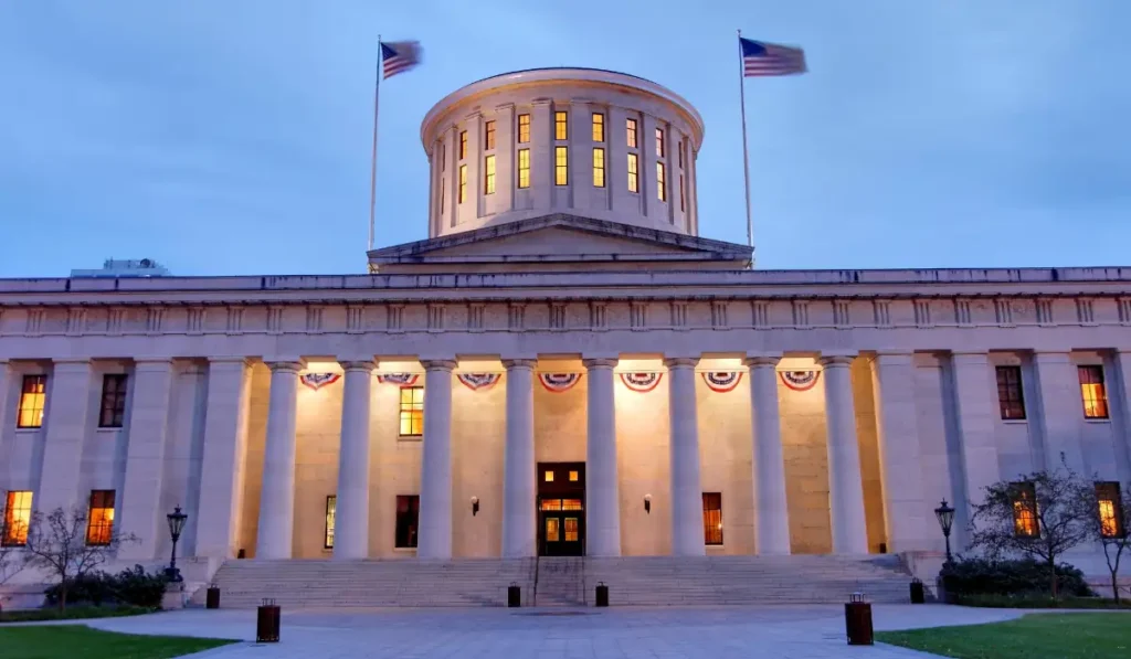 The image shows the Ohio Statehouse, a large building with a distinctive rotunda and two American flags flying atop. The neoclassical architecture is highlighted by warm lights that illuminate the columns and windows against the evening sky.
