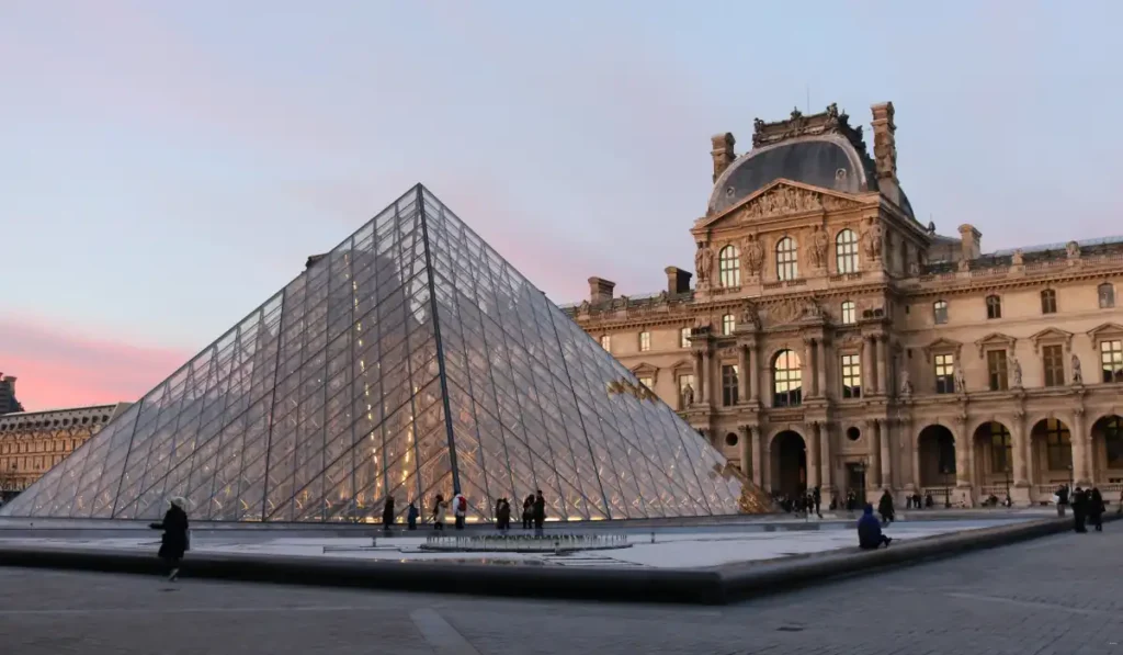 Glass pyramid entrance to the Louvre Museum at sunset, with the historic museum building in the background and people walking around the courtyard.
