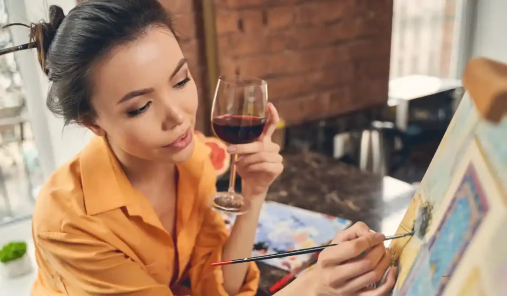 A person in an orange shirt holds a glass of red wine while painting on a canvas in a cozy room with brick walls. They appear focused on their artwork.
