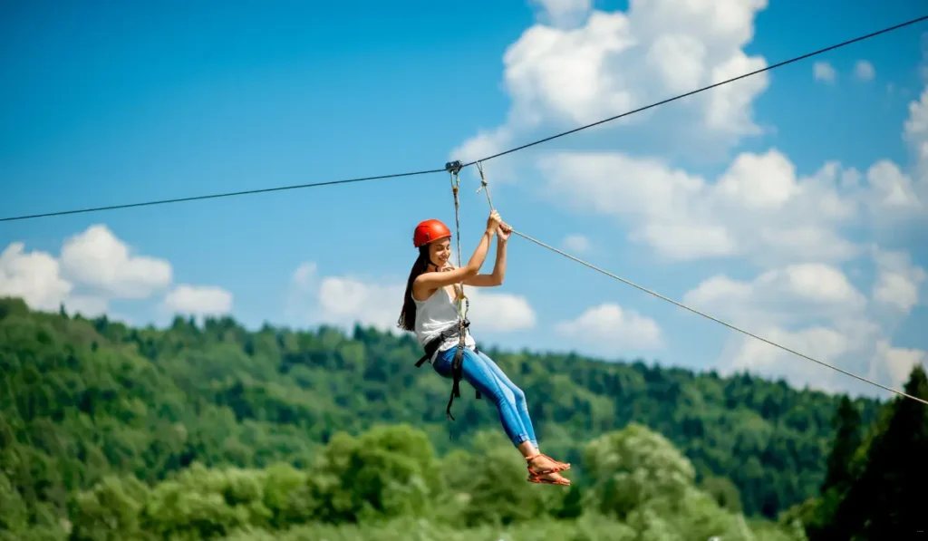 A girl wearing a red helmet and harness zip-lining over a lush green forest under a bright blue sky with scattered clouds.
