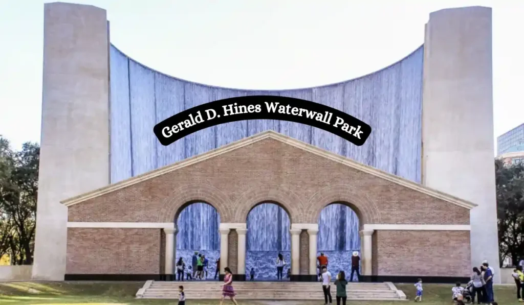 A large architectural structure with cascading waterfalls is labeled Gerald D. Hines Waterwall Park. People gather around the arches and brick facade, enjoying the outdoor setting. Trees frame the background.
