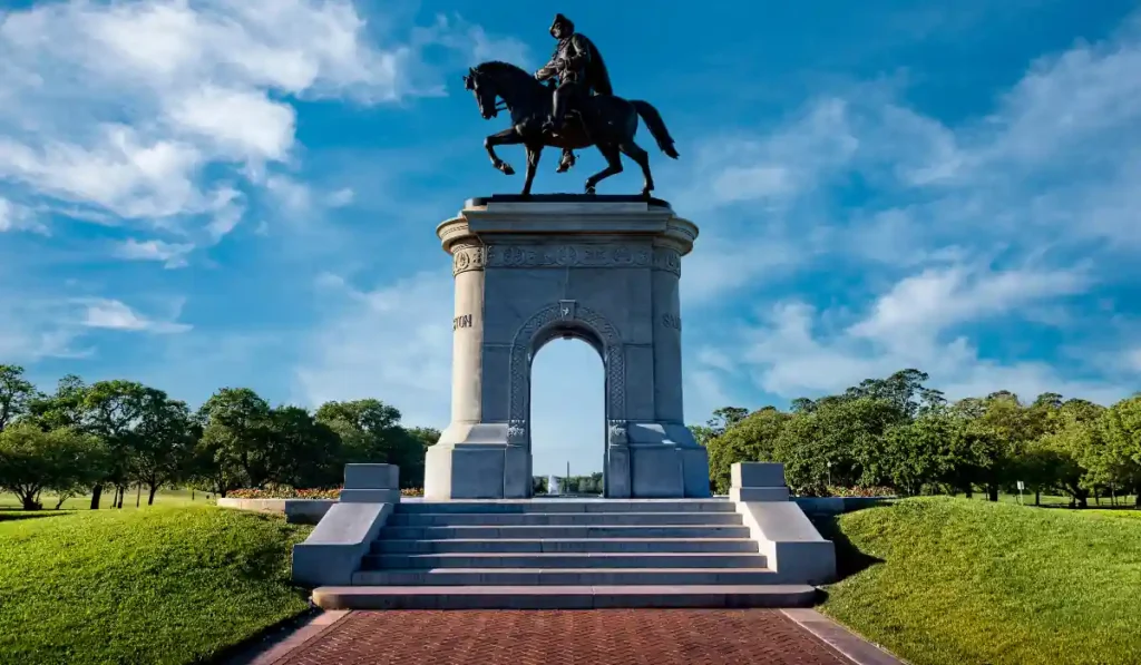 A bronze equestrian statue of a uniformed rider on a horse stands atop a large stone pedestal. The statue is framed by a clear blue sky and surrounded by green grass and trees. Stone steps lead up to the base of the monument.
