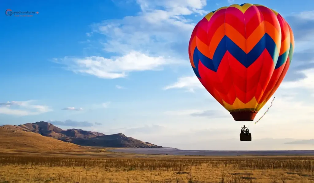 A colorful hot air balloon with a vibrant zigzag pattern floats above a vast, open landscape with dry grass and distant hills under a clear blue sky.
