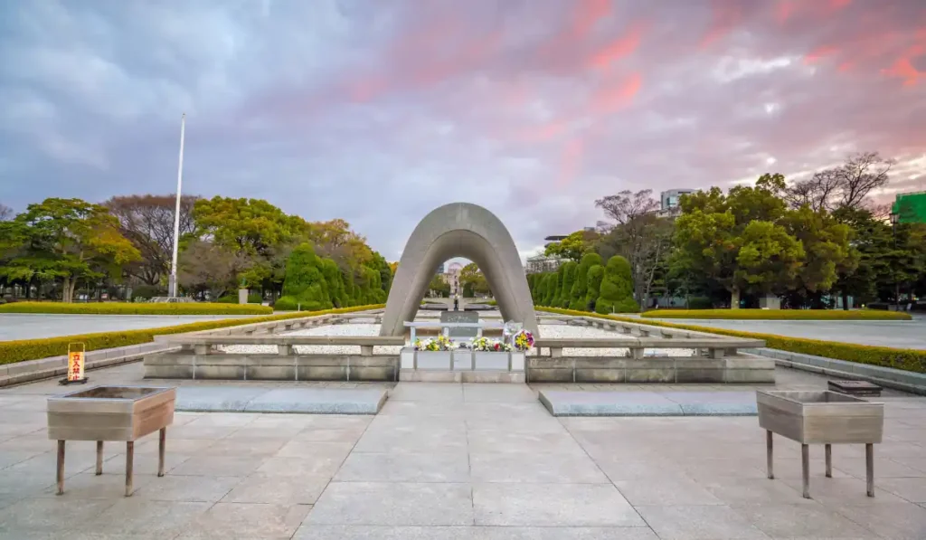 A peaceful scene of the Hiroshima Peace Memorial Park at sunset, featuring the Memorial Cenotaph surrounded by green trees. The sky is painted with soft shades of pink and purple.
