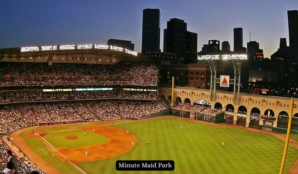 A baseball game at Minute Maid Park during sunset. The stadium is filled with spectators, and players are on the field. The city skyline is visible in the background, and the outfield wall displays advertisements.
