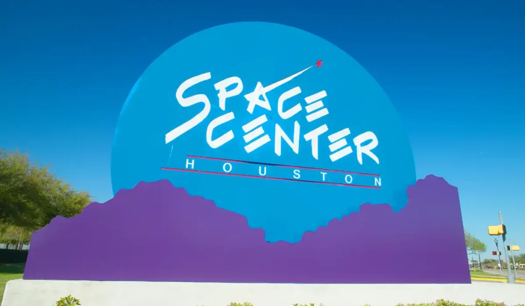 A large blue and purple sign for Space Center Houston against a clear blue sky. The words Space Center Houston are displayed in bold white letters, with a red arrow motif. Trees and a streetlamp are visible in the background.
