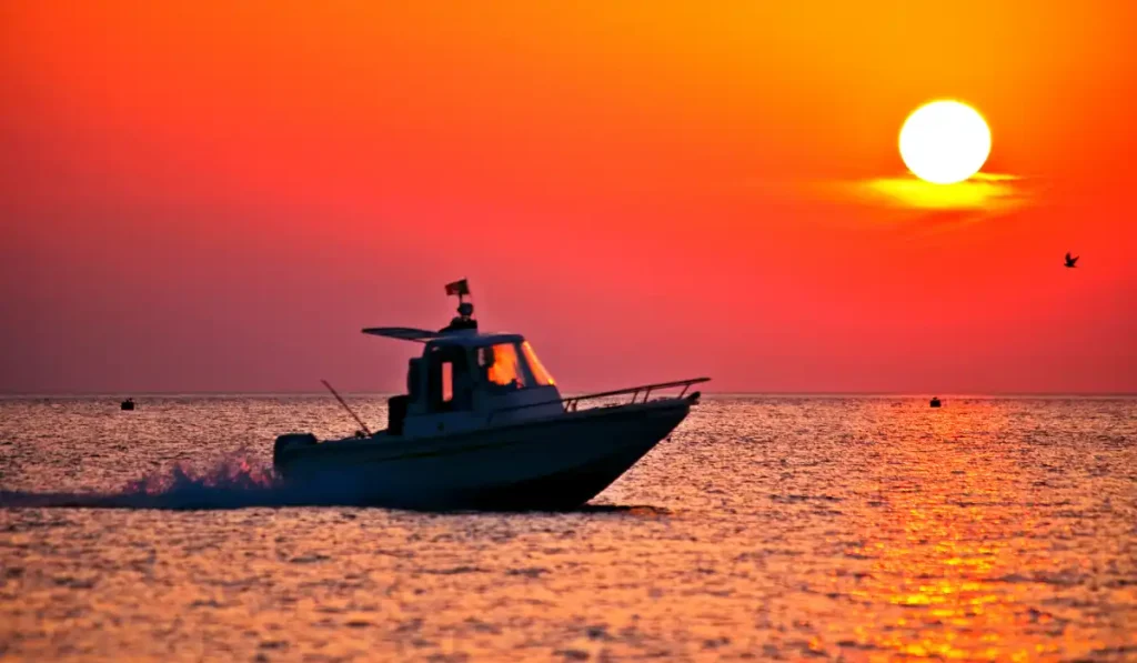 A silhouette of a boat speeds across the water at sunset, with the sun setting in a vibrant orange and red sky. A small bird is flying to the right, and the ocean reflects the warm colors of the sky.
