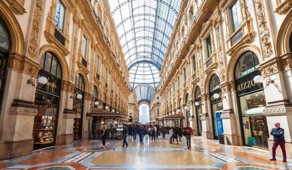 A bustling shopping arcade with a glass-vaulted roof, ornate architecture, and an intricate tiled floor. People are walking and shopping, surrounded by elegant storefronts and soft lighting.
