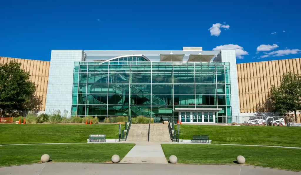 A modern building with a large glass facade reflecting the blue sky and white clouds. It is surrounded by green lawns and trees, with steps leading to the entrance. The structure features a blend of glass and light-colored materials.
