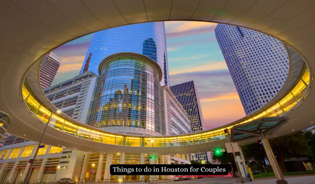 A view of downtown Houston skyscrapers seen through a circular architectural structure at sunset. The sky is vibrant with pink, orange, and blue hues. A caption reads, Things to do in Houston for Couples.
