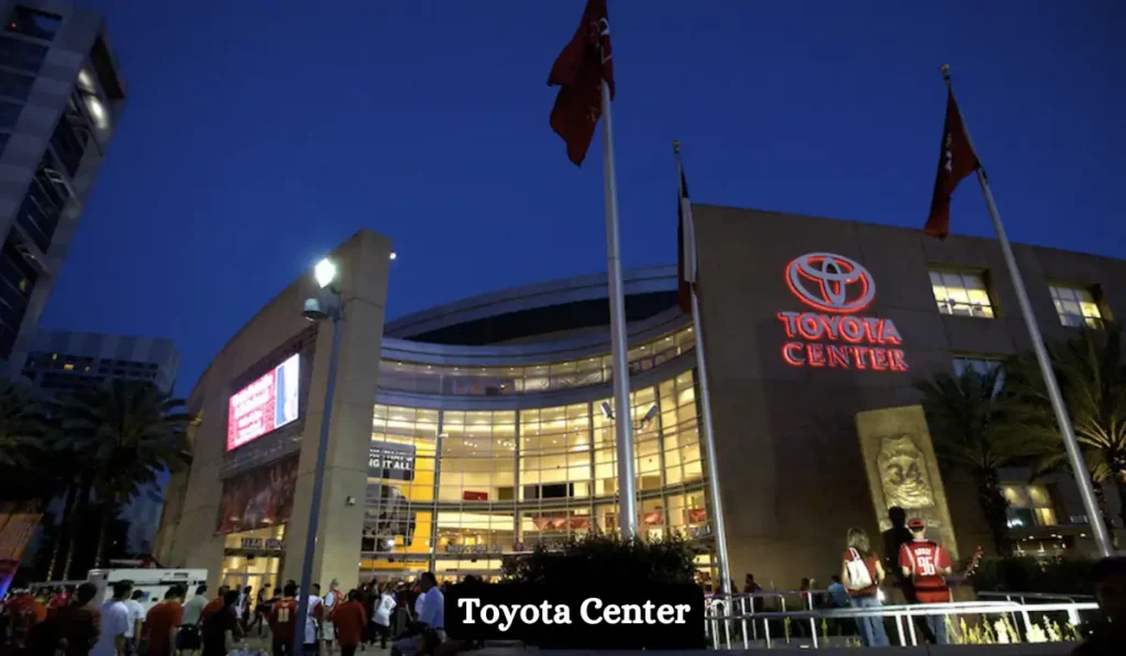 Night view of Toyota Center with its brightly lit glass facade. American flags and people in sports jerseys are visible in the foreground.
