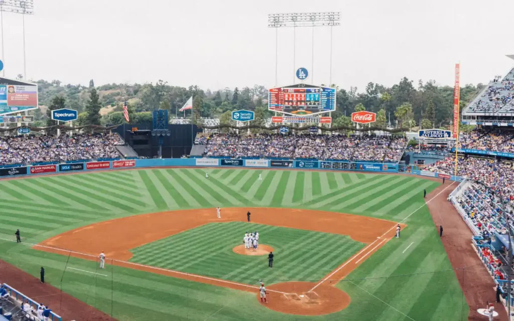 A baseball game in progress at a large stadium. The field shows neatly mowed grass with a dirt infield. Players are gathered on the pitchers mound, surrounded by an audience in the stands under a cloudy sky. Advertising banners are visible.