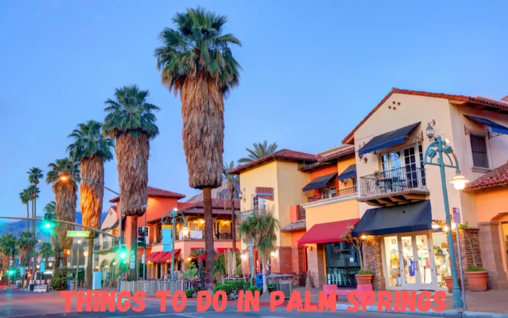 A colorful street scene in Palm Springs features tall palm trees, vibrant storefronts, and warm Mediterranean-style architecture. The text THINGS TO DO IN PALM SPRINGS is prominently displayed in red at the bottom of the image.