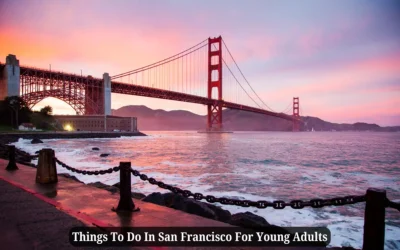 The image shows the Golden Gate Bridge at sunset, casting a warm glow over the water. The foreground features a rocky shoreline and a chain-link fence. Text at the bottom reads, Things To Do In San Francisco For Young Adults.